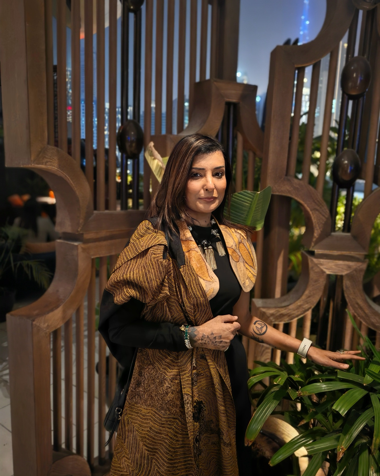 Pallavi in front of a carved wooden screen, city lights behind